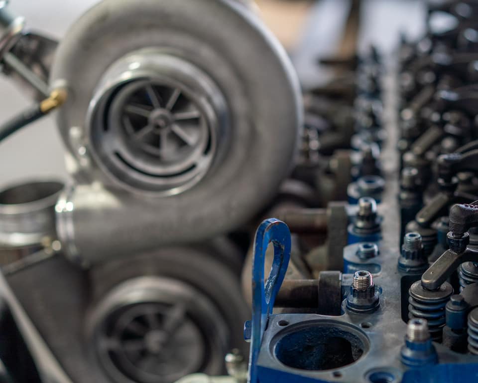 Close-up of an automotive engine component, featuring a silver turbocharger on the left and a blue-painted engine block with metal bolts and springs on the right.