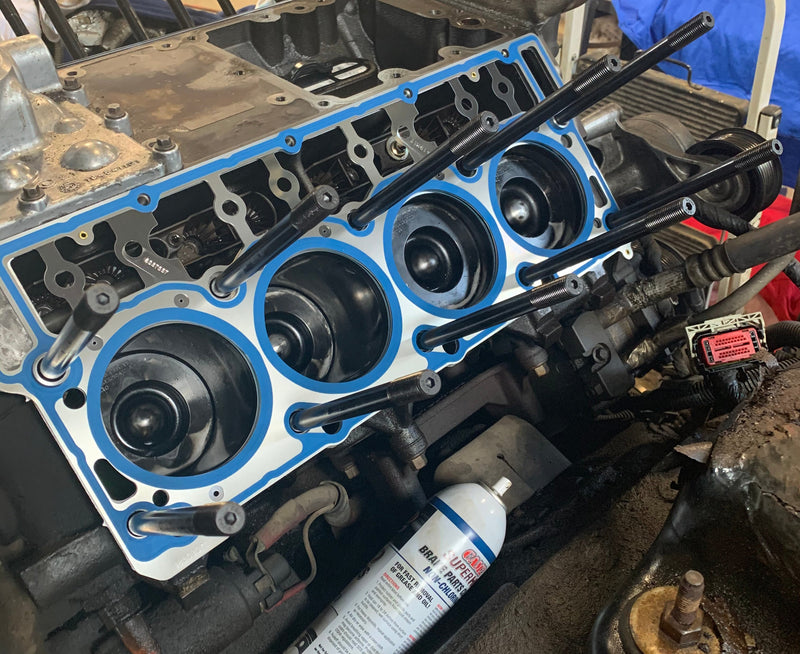 A close-up of a partially disassembled engine block in a workshop setting, showing the blue and black valve cover gasket area, black intake manifold, and surrounding mechanical components.