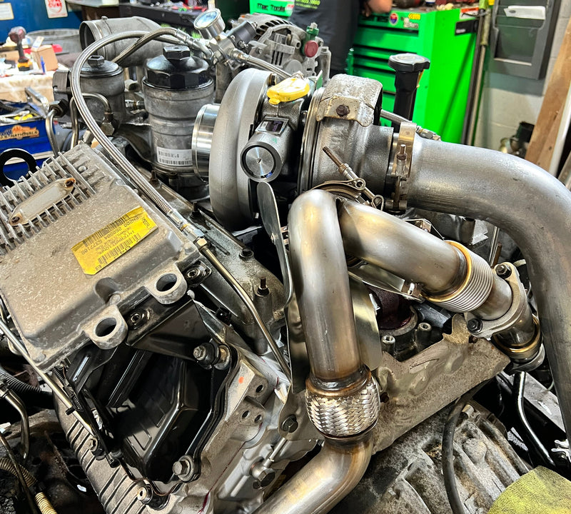 A close-up of a high-performance automotive engine with polished stainless steel intake pipes and turbos, set in a workshop with a mechanic working in the background.