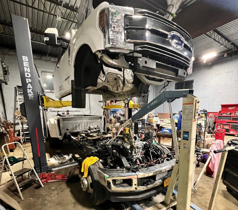 A white pickup truck is shown in a auto repair shop with its front end raised on a lift. The front bumper, grille, headlights, and hood are removed, exposing the engine bay.